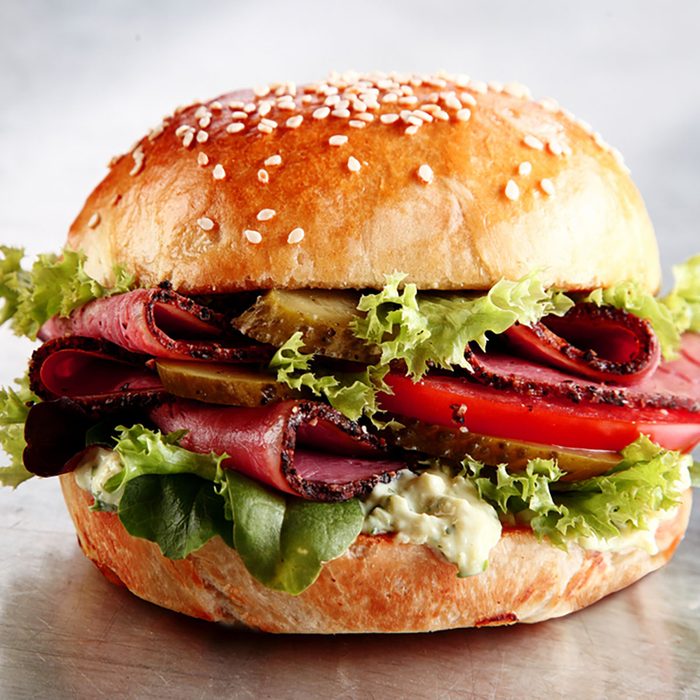 Delicious sesame bun with sliced roast beef or pastrami with lettuce, tomato, gherkin, and mayo on a silver counter in a cafeteria or restaurant