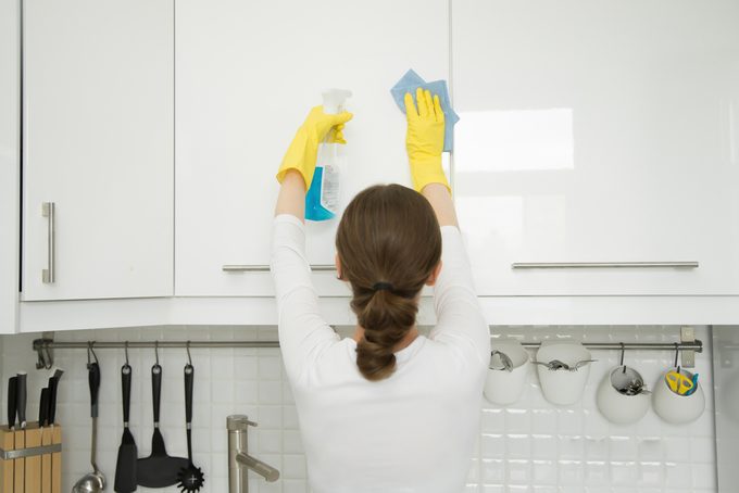 Rear view at an attractive young woman cleaning a surface of white kitchen wall cabinet, wearing rubber protective yellow gloves, with rag and spray bottle detergent. Home, housekeeping concept