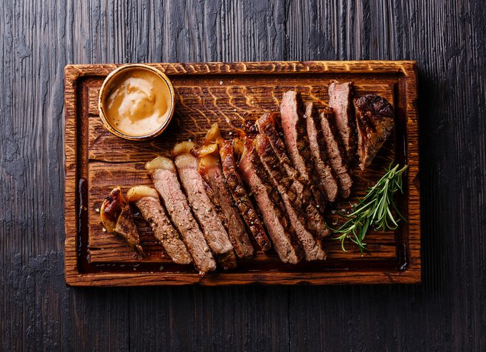 Sliced grilled well done Striploin steak with Pepper sauce on cutting board on burned black wooden background