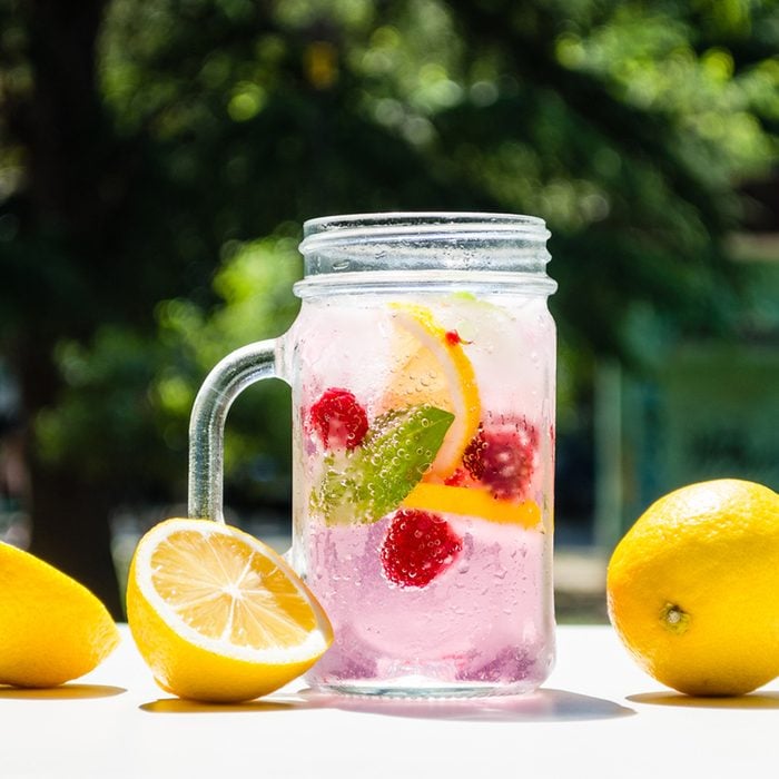 Healthy detox water served in a mason jar with ice lemon raspberries mint leaf bubbles and surrounded with green nature and trees into the background