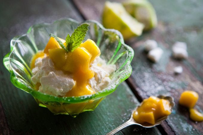 Fresh mango and lime syllabub served in a dainty emerald green glass dessert dish garnished with mint and icing sugar on distressed green wooden work surface