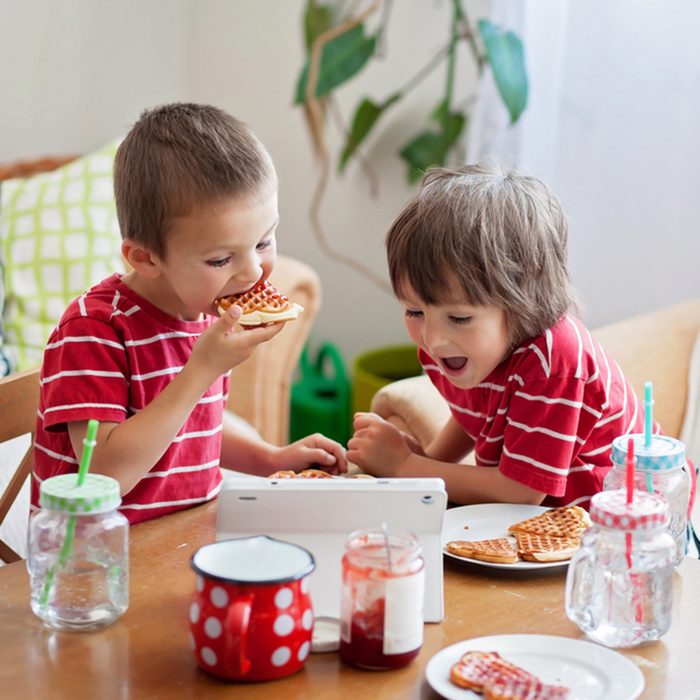 Two happy kids, two brothers, having healthy breakfast sitting at wooden table in sunny kitchen, eating waffles and watching cartoon on tablet