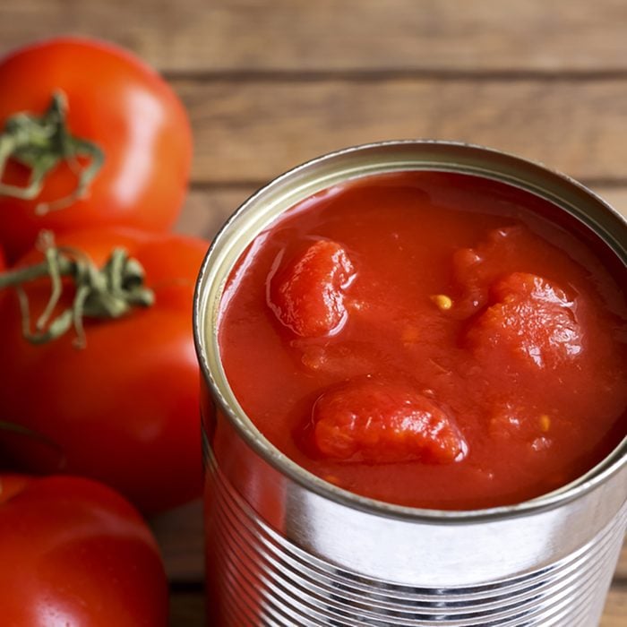 Open tin of chopped tomatoes with whole fresh unfocused tomatoes behind. Wood surface
