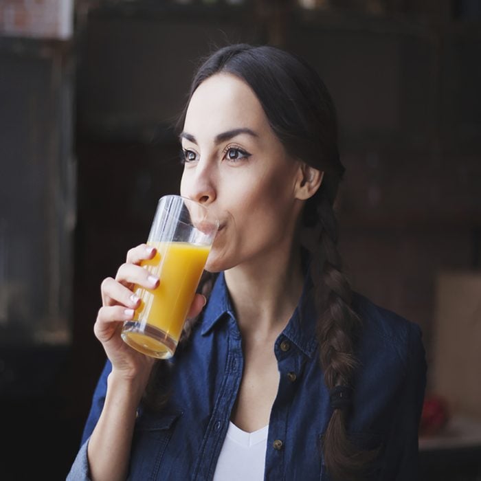 Portrait of very beautiful young brunette girl in jeans shirt on a kitchen at home. The girl drinks orange juice from transparent glass.