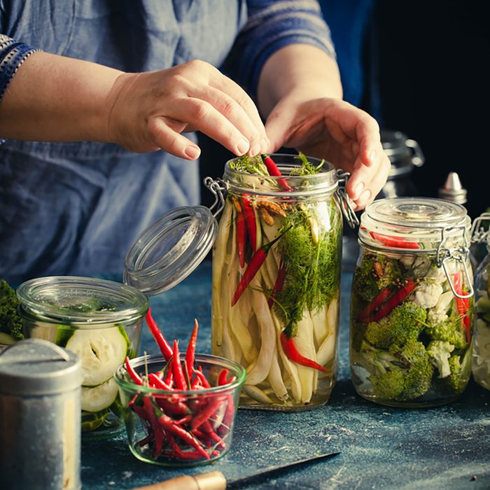 Canning green beans with red hot chili peppers in glass jars with pickled cabbage cauliflower broccoli sour preserved hands add herbs fermented process.