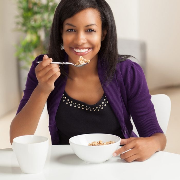 Beautiful young black female sitting at dining room table eating breakfast and drinking coffee.
