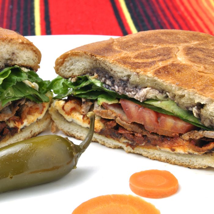 Closeup of halved Mexican torta sandwich with toasted bun and jalapeno pepper on plate over colorful tablecloth