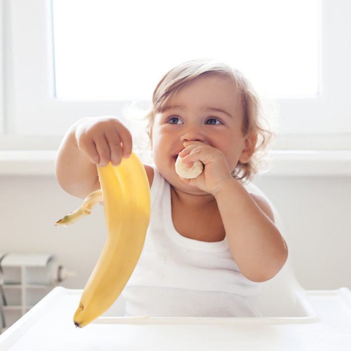 4 years old sitting on high children chair and eating fruit alone in white kitchen