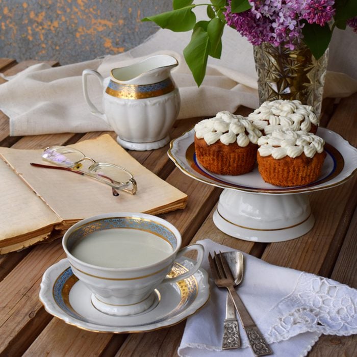 Romantic composition of bouquet white and purple lilacs, cupcakes with curd cream, cups tea with milk, old book on wooden background.