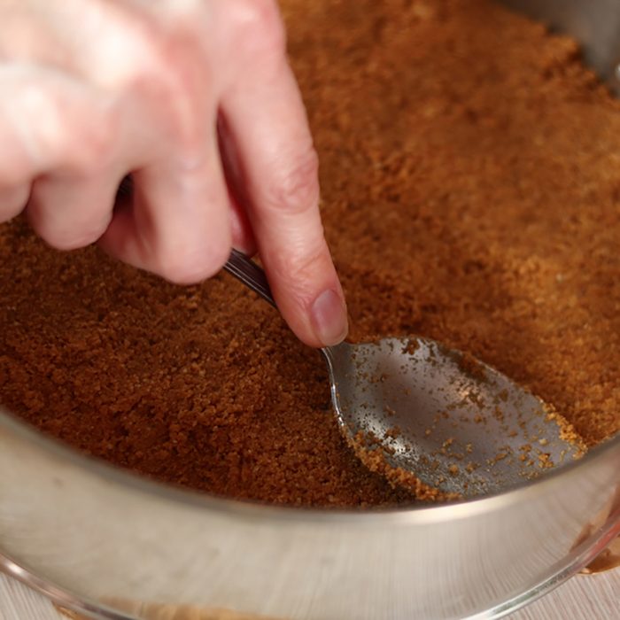 Leveling ground cookies mixture with spoon in baking pan. Making frozen strawberry cheesecake series.