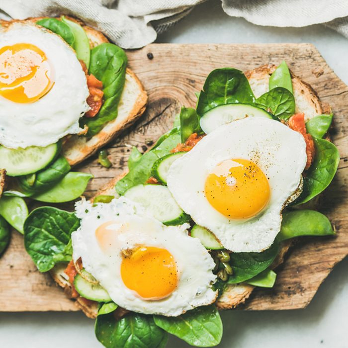 Bread toasts with fried eggs and fresh vegetables on rustic wooden board over grey marble background, top view.