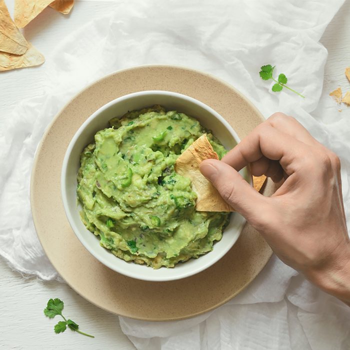 Guacamole freshly cooked and served in a bowl, overhead view on woman hand is picking some guacamole dip with pita chip