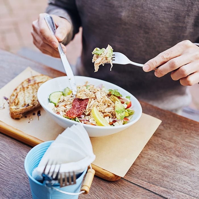 Cropped image of a man on his lunch break eating a fresh and healthy salad with chicken, avocado, sundried tomatoes and fresh sliced baguette on the side while sitting at a wooden table.