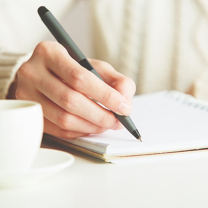 Woman writing in spiral notepad placed on bright desktop with coffee cup.