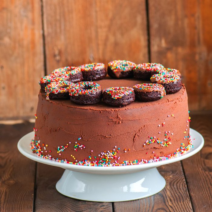 Close up of homemade rich chocolate cake with chocolate ganache frosting decorated with baked doughnuts with confetti glaze on a white cake stand on a wooden background.