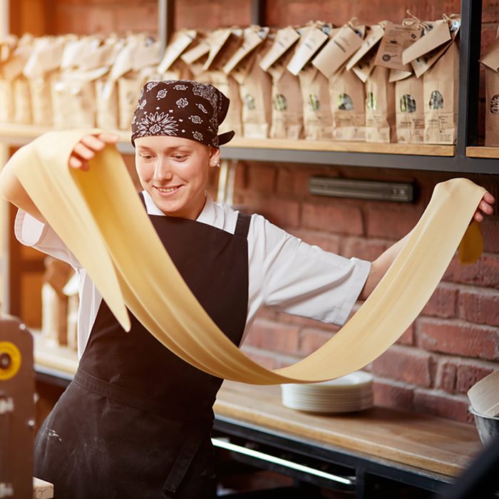 Cheerful young woman rolling a knead through pasta machine and smiling. 