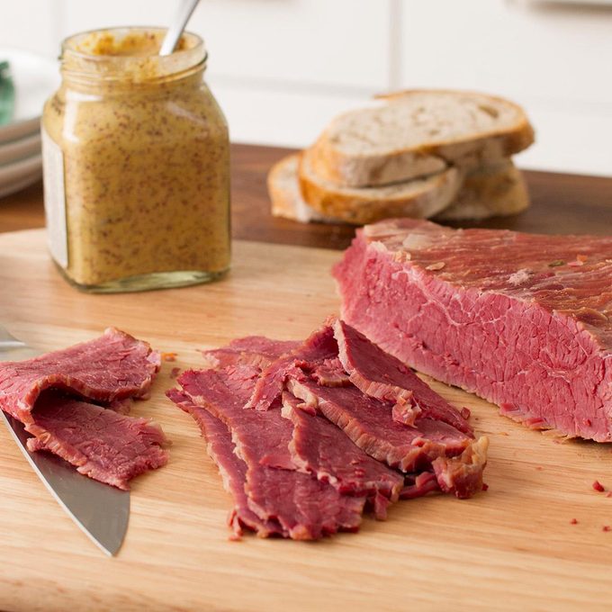 A piece of sliced corned beef brisket on a cutting board. With a jar of mustard and rye bread in the background.