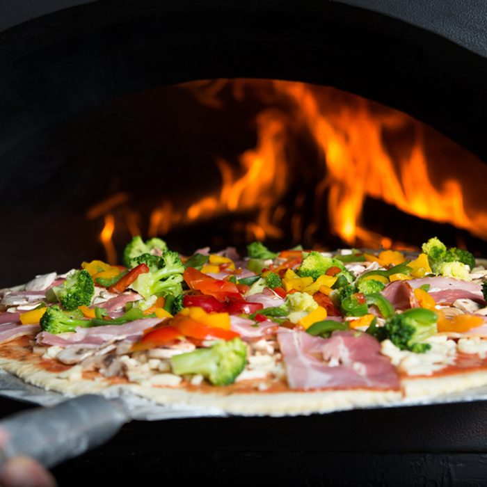 The cook puts pizza bread into wood-burning stove.
