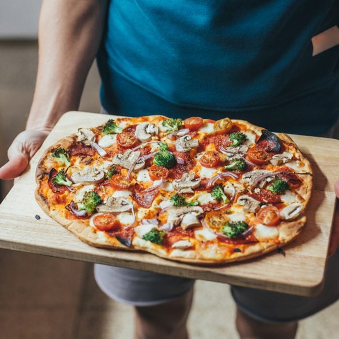 cook holds wooden tray or board with homemade organic flatbread pizza, covered with vegetables, veggies and cheese