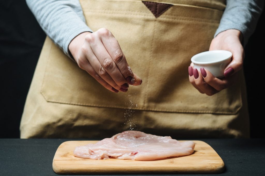 Woman hand sprinkle raw chicken fillet by condiment.