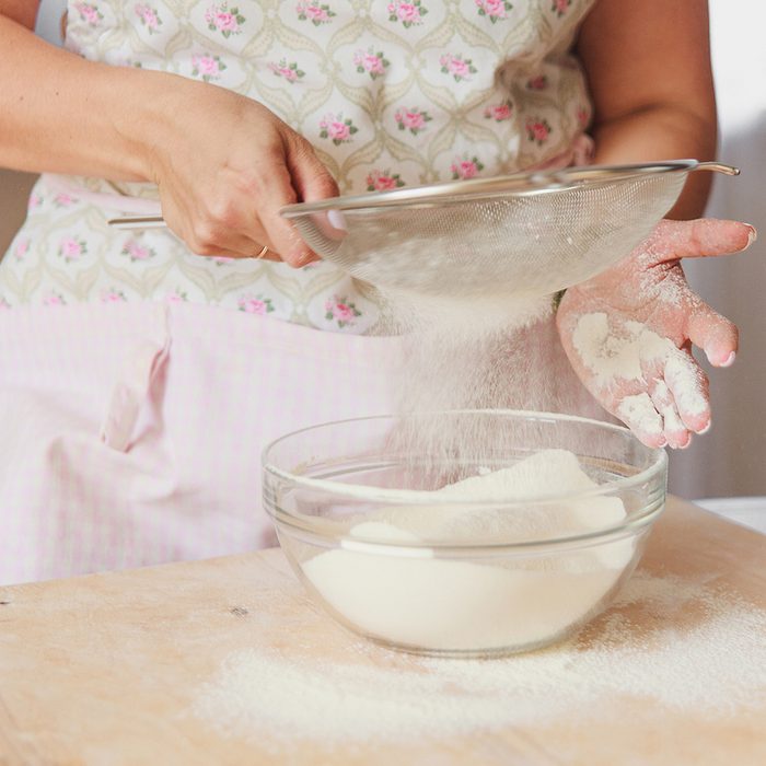 Aged female hands sifting flour by sieve in glass bowl.