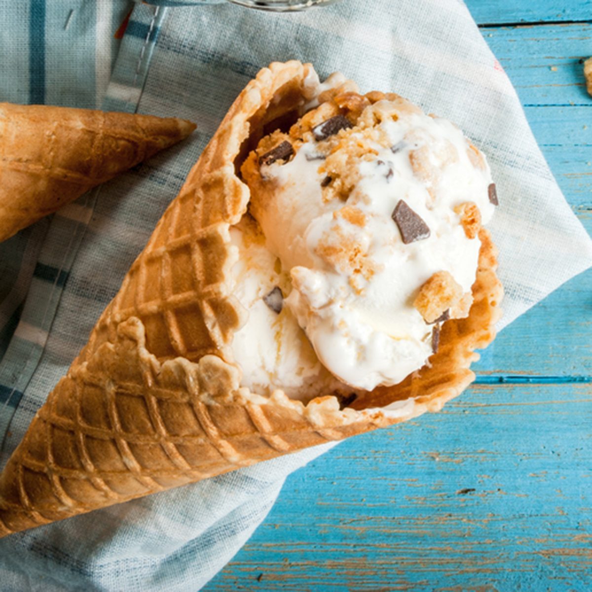 Traditional waffle cones for ice cream and a spoon for it on blue wooden table. Summer, open space, bright sun Wafers and chocolate chips in a frame on the table One of cones filled with ice cream; Shutterstock ID 518542783; Job (TFH, TOH, RD, BNB, CWM, CM): Taste of Home