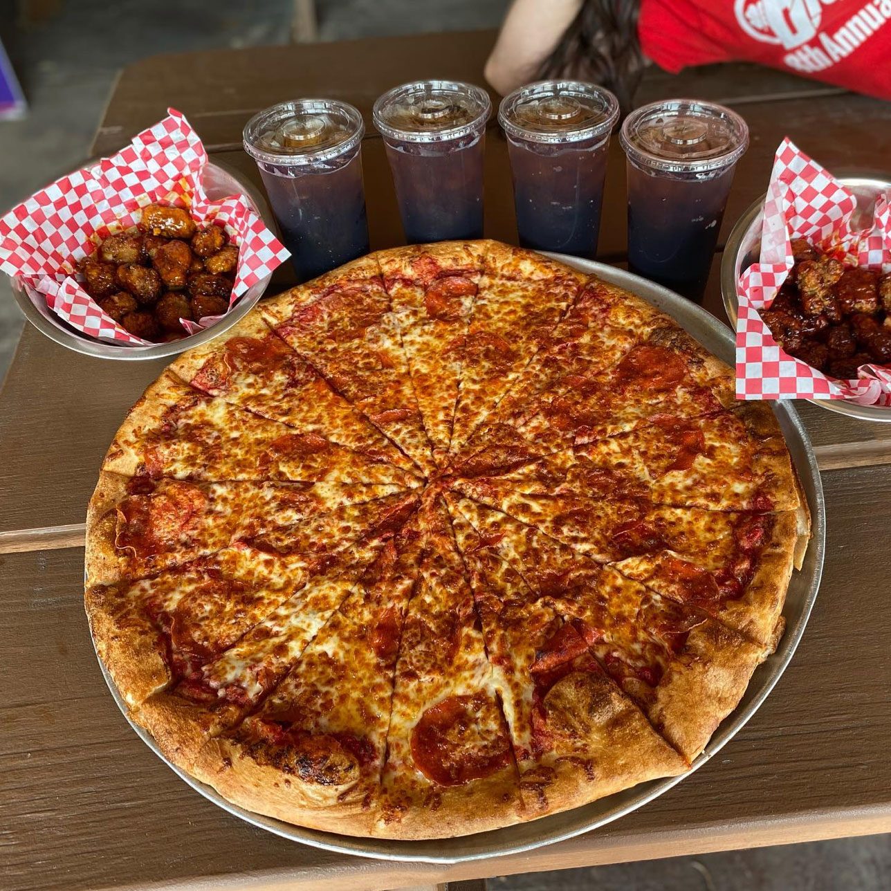 cheese and pepperoni pizza with two baskets of wings and four cups of soda in the background