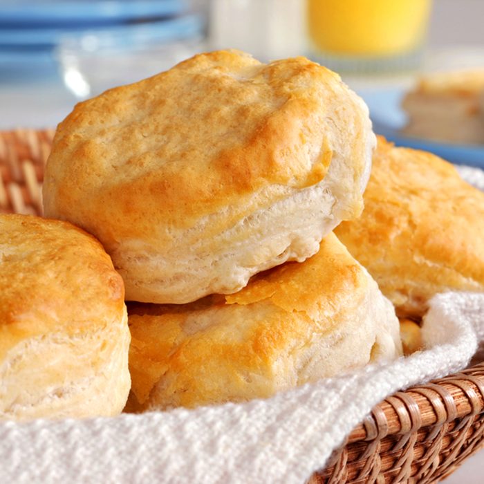 Basket of freshly baked biscuits with orange juice and tableware in background.