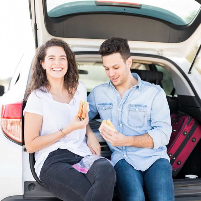 Beautiful young woman sitting in car boot with her boyfriend eating sandwiches, lunch break on road trip