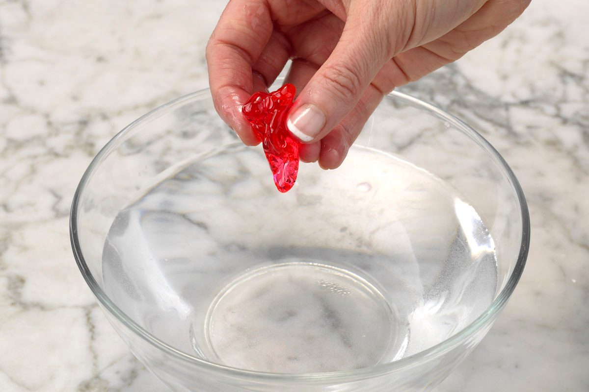 Testing red homemade candy using the cold water test. The mixture forms a ball when dipped in cold water and remains hard yet pliable ball after being removed, which means it's in hard-ball stage.