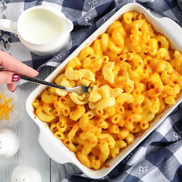 woman's hand is holding a fork with small portion of Mac and cheese or elbow pasta baked with cream and sharp cheddar cheese sauce in dish,