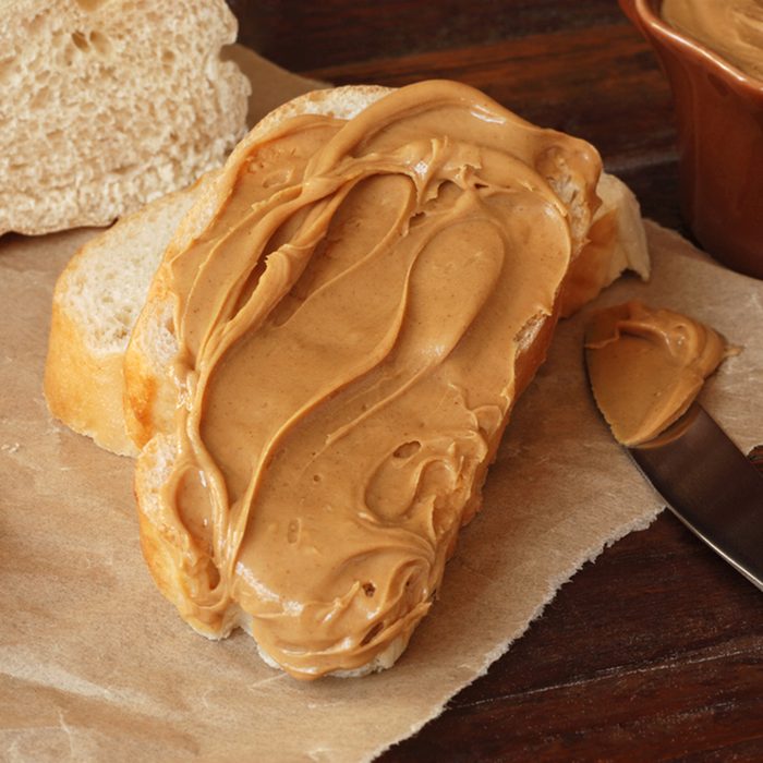 Creamy swirls of peanut butter on freshly baked italian bread with knife and crumpled parchment paper over rustic wood cutting board.