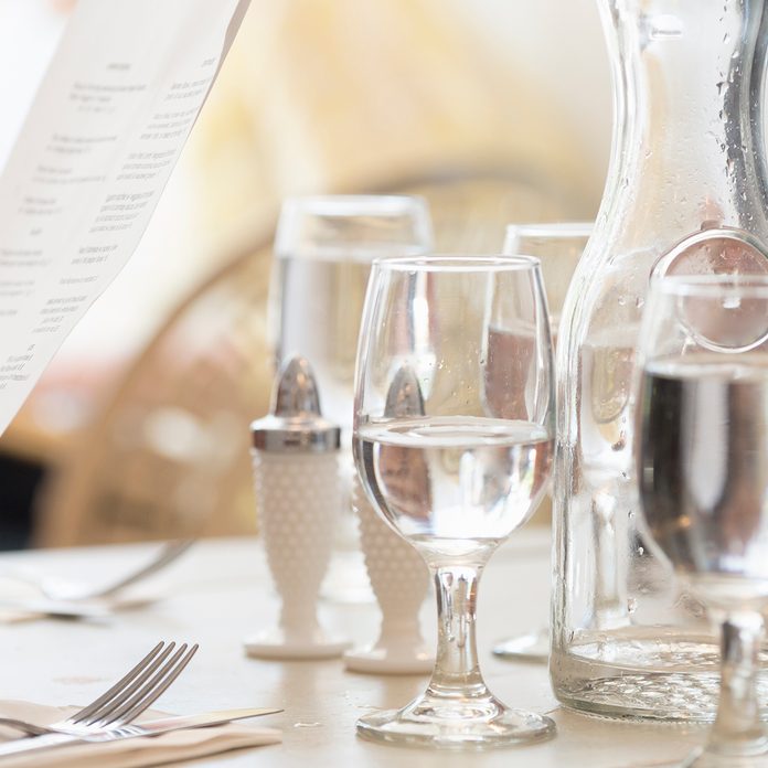 Close Up Of Wine And Water Glasses And Place Settings At A Table In A Restaurant. A Persons Hand Holding The Menu.