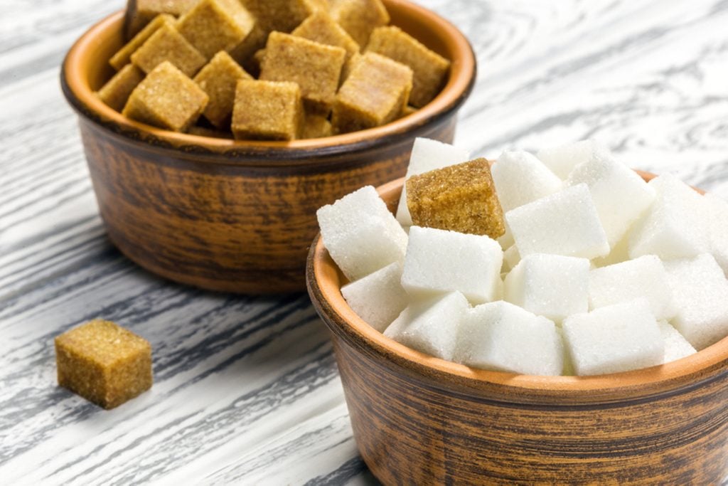 White refined sugar and brown unrefined sugar cubes in ceramic bowls on a light wooden table.