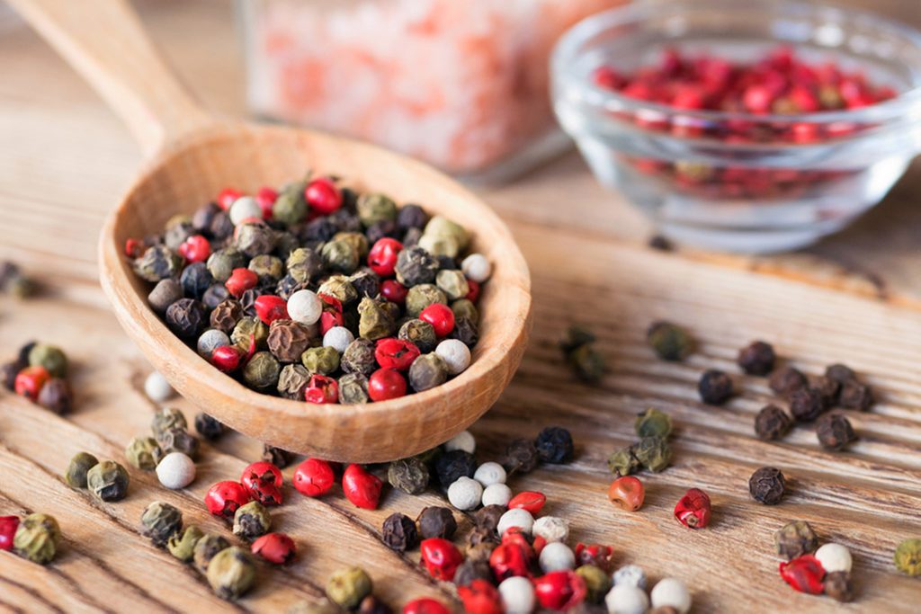 Mix of peppercorns in wooden spoon on rustic background with pink salt in glass jar.