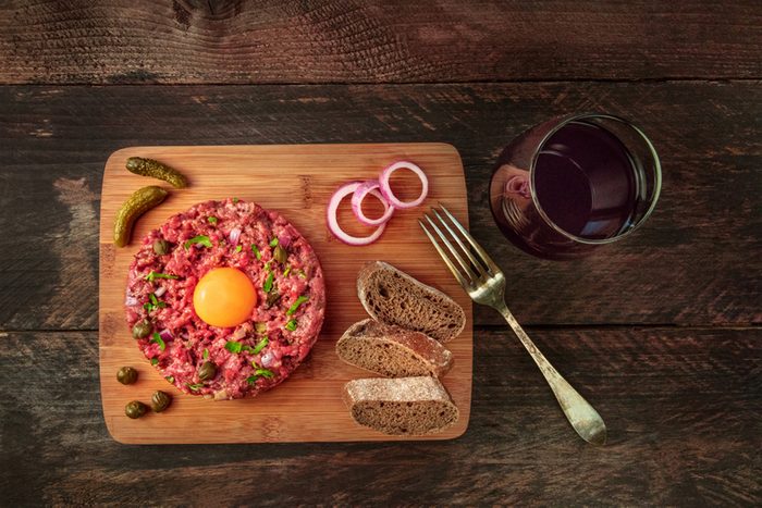 A photo of a steak tartare with a raw egg yolk, gherkins, capers, rye bread, purple onions, a glass of red wine, and a fork, shot from above on a dark rustic texture