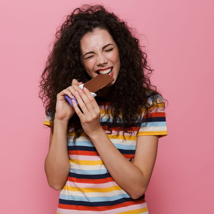 Photo of brunette woman 20s with curly hair eating chocolate bar isolated over pink background