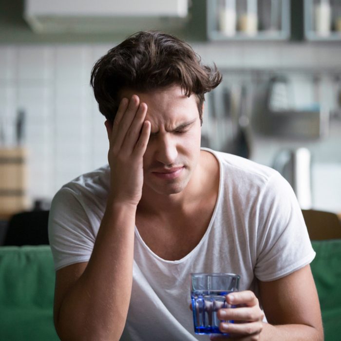 Young man suffering from strong headache or migraine sitting with glass of water in the kitchen