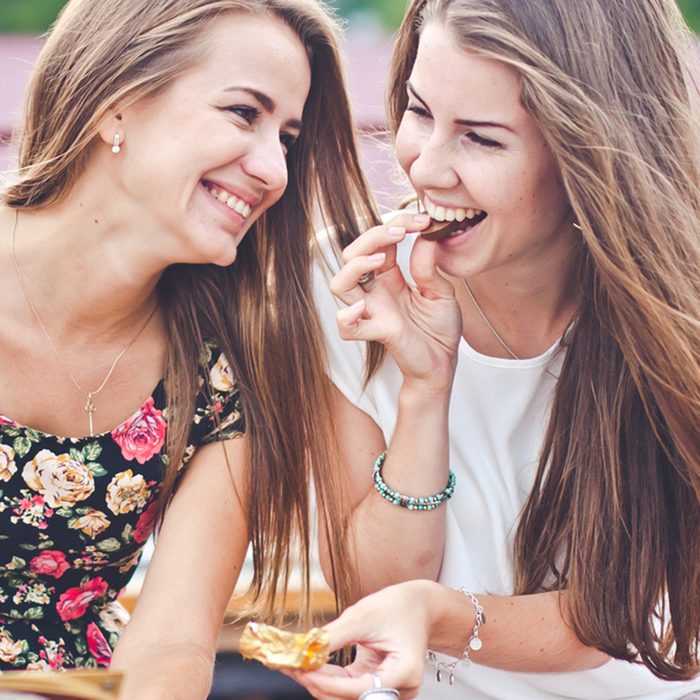 Two young female friends laugh and eat chocolate outdoors 