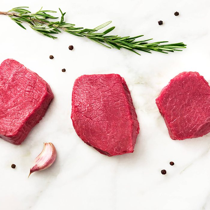 Three slices of raw meat, beef fillet, shot from above on a white marble table with a sprig of rosemary, garlic cloves, salt, and pepper