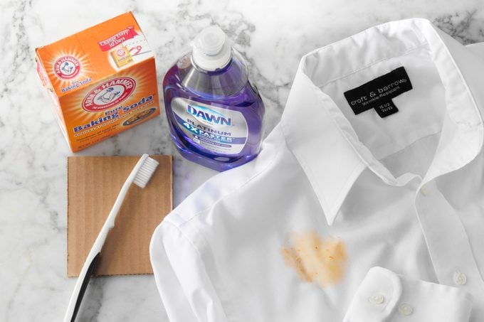White Shirt Stained with pizza grease next to stain cleaning supplies: Baking Soda, Dawn dish soap, small piece of cardboard, And a Tooth Brush