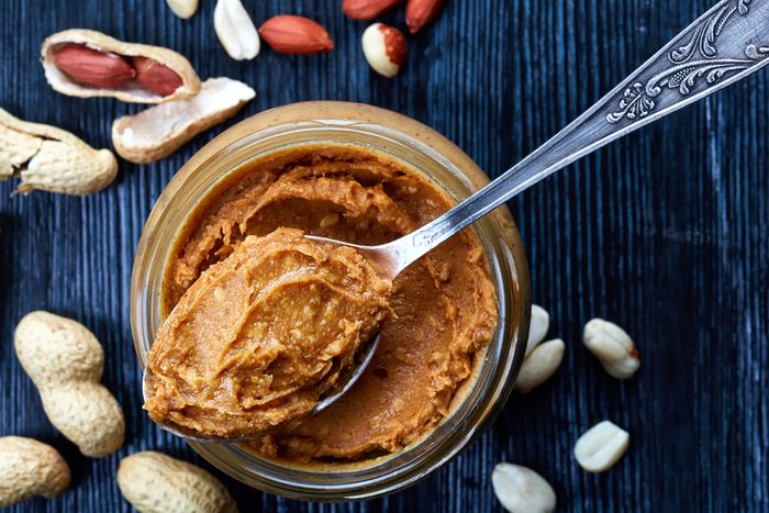 Jar and spoon of peanut butter and peanuts on dark wooden background