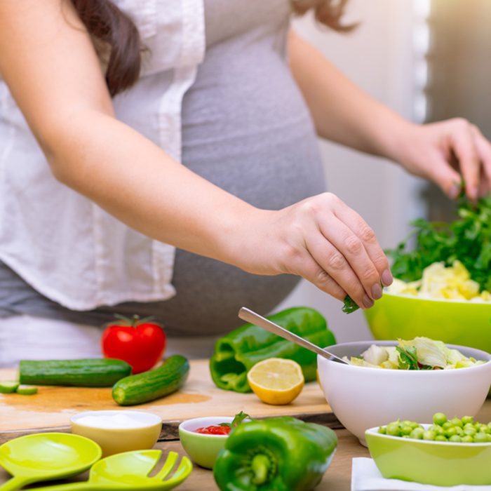 Happy pregnant woman cooking at home, doing fresh green salad, eating many different vegetables during pregnancy
