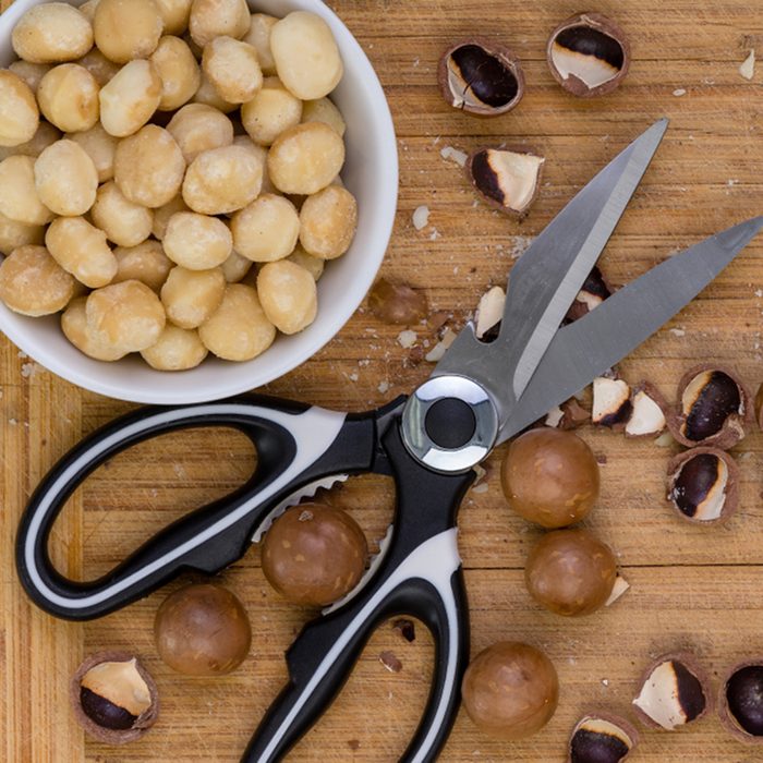 Pair of kitchen shears or scissors with shelled, cracked and whole macadamia nuts on a wooden bamboo cutting board viewed close up from above; Shutterstock ID 1012493167