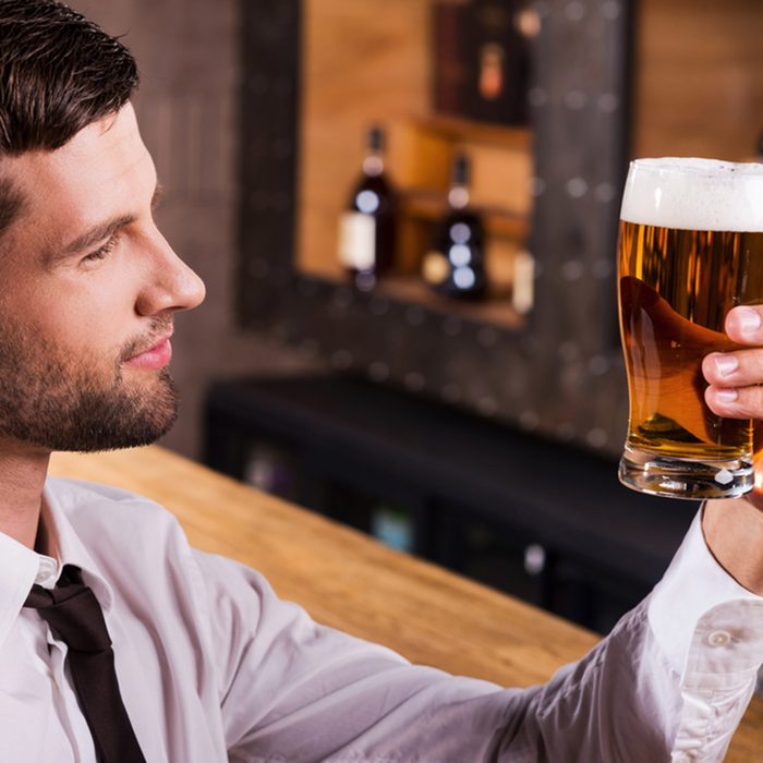 Side view of handsome young man in shirt and tie examining glass with beer and smiling while sitting at the bar counter