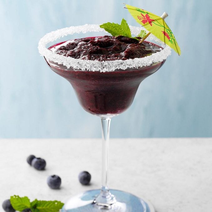 A homemade blueberry-mint frozen margarita in a glass with a salted rim and a decorative umbrella.