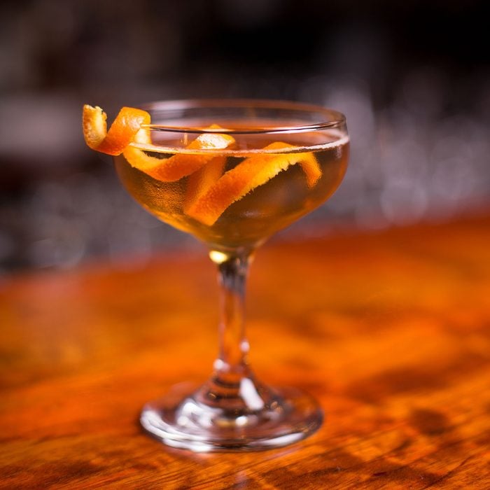 Low angle closeup of gourmet old fashion martini drink in champagne saucer cocktail glass filled with bourbon and brandy garnished by delicately spiral orange peel on red cherry wood grain countertop