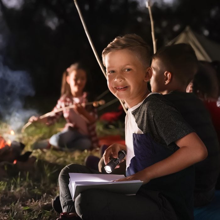 Little boy with book and flashlight near bonfire at night. 