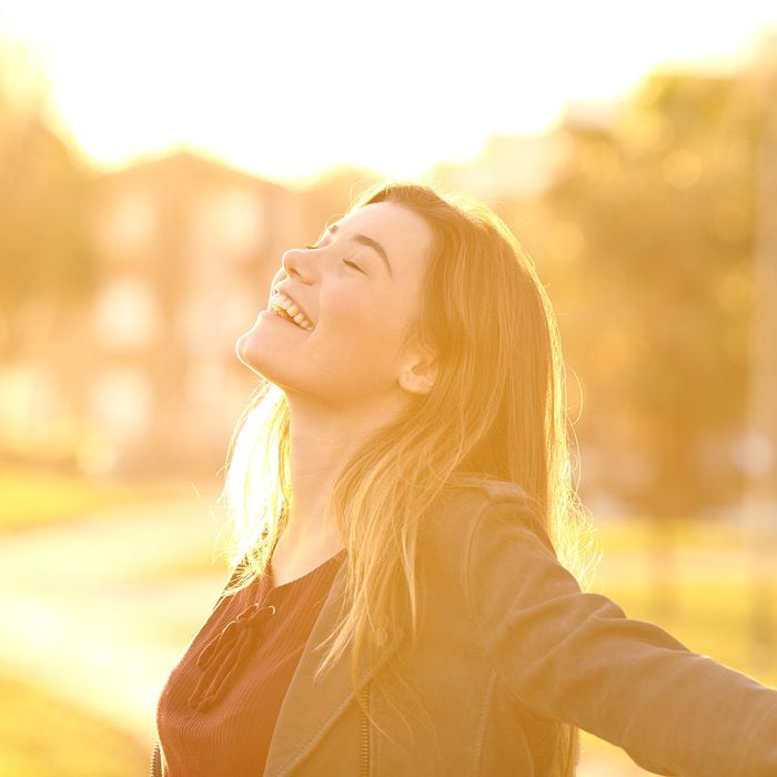 Back light portrait of a happy single teen girl breathing fresh air at sunset in a park with a warm yellow light and urban background