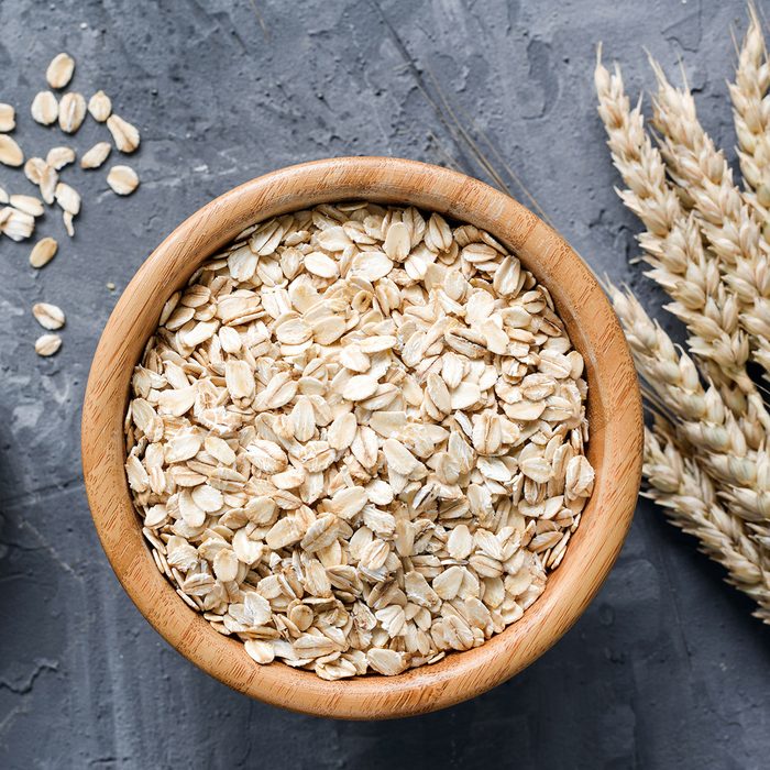 Rolled oats or oat flakes in wooden bowl and golden wheat ears on stone background.